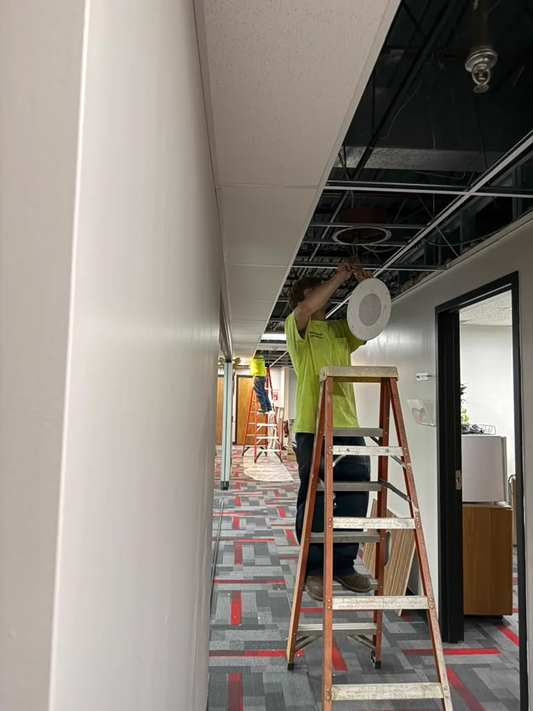 Excel Facility technician on a ladder installing a ceiling light fixture in an office corridor
